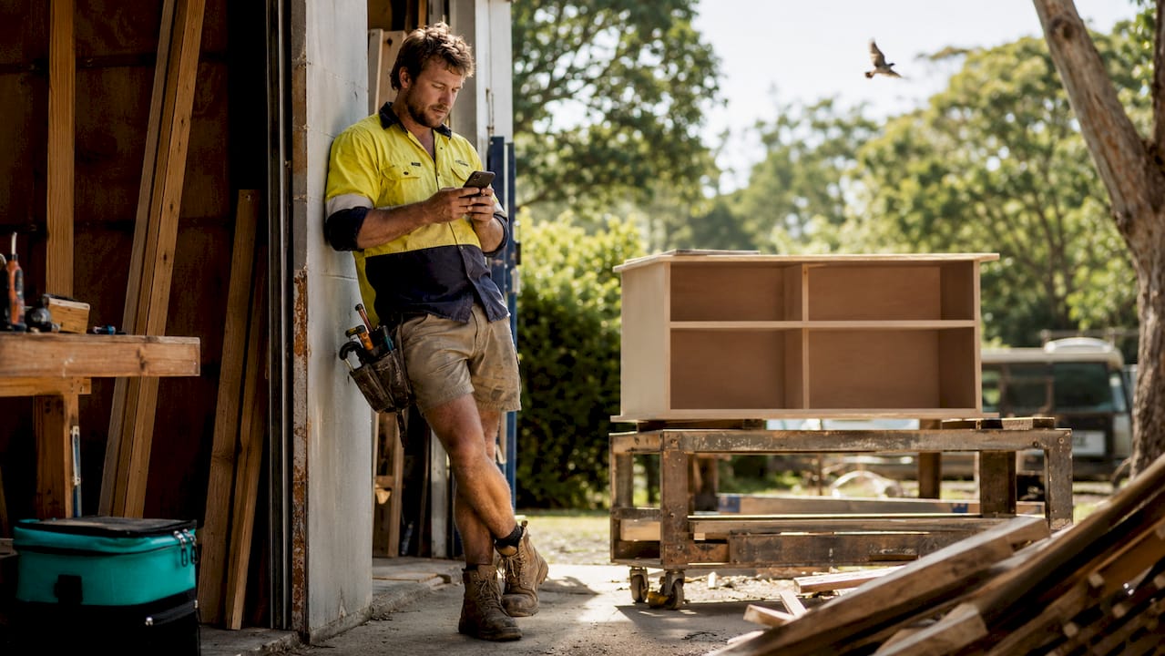 1782823877916_Tradie-scrolling-social-media-on-phone-in-workshop Tradie scrolling social media on phone in workshop